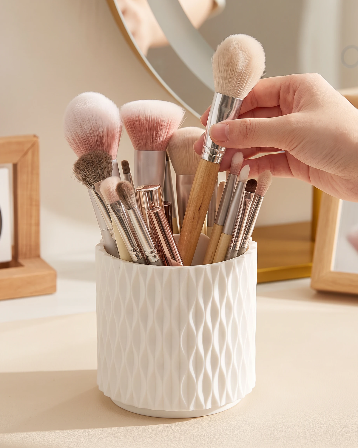 A hand holding a makeup brush with a light wood handle and beige bristles, standing beside a white textured cup filled with various makeup brushes on a vanity table.