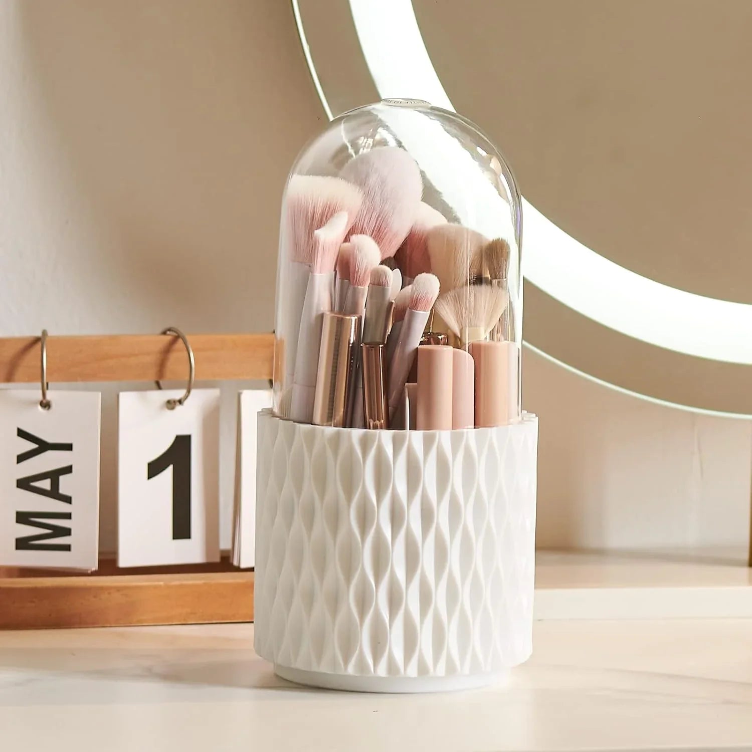 Makeup brushes in white textured holder with clear lid on vanity near May 1 calendar and mirror